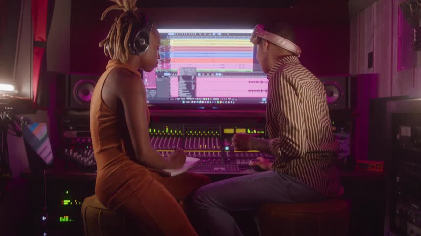 Two young women one black and one caucasian sitting in a music studio wearing headsets nodding their heads to the music theyre listening to while one adjusts the sound levels on the mixer
