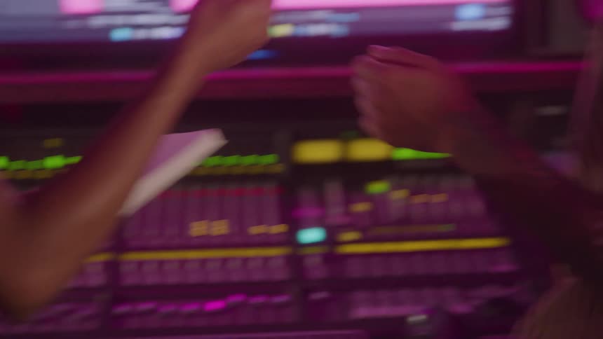 Two young women in a music studio both wearing headsets standing in front of a computer screen they are enjoying the music while discussing with one holding a pen and a notebook taking down notes