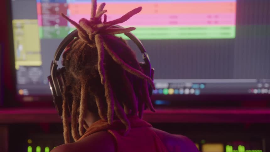 Young black woman with braided hair wearing headphones sitting behind a computer monitor in a music studio nodding her head in rhythm to the song shes listening to