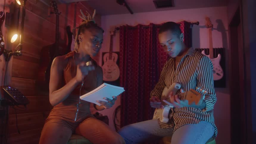 Two young women are collaborating on a song in a music studio with one woman playing an electric guitar and the other holding a notebook and gesturing enthusiastically as they talk