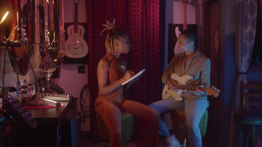 Two young women sitting on stools in a music studio one playing a guitar while the other writes in a notebook with a pen the room is filled with various musical instruments