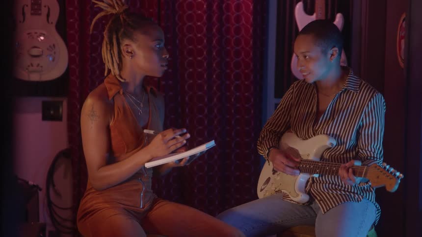 Two women are collaborating in a dimly lit room with musical instruments one playing a guitar and the other writing in a notebook