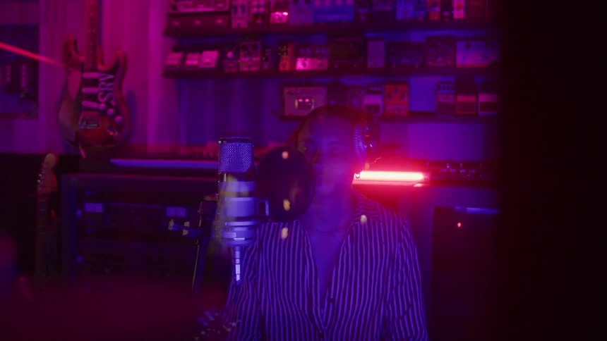 A man singing into a microphone in a studio while bobbing his head and the room is lit by a neon pink light