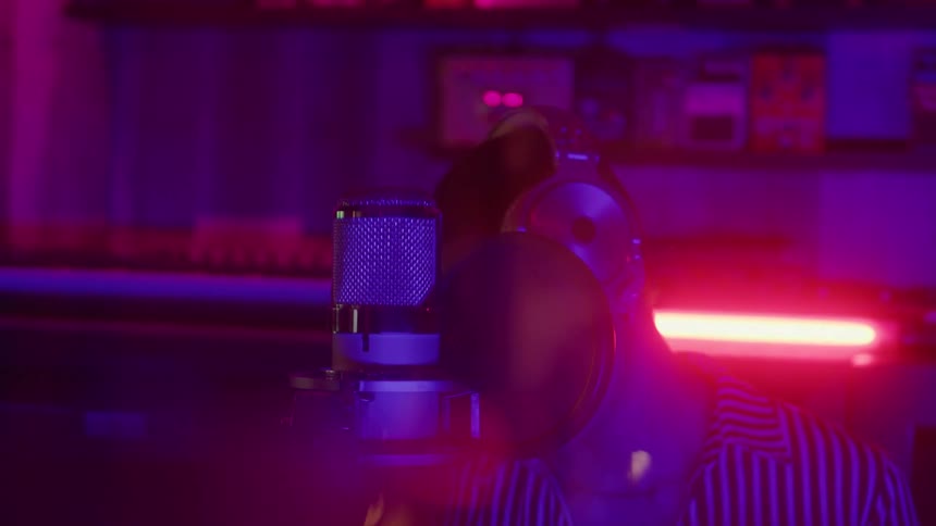 The face of a man who is singing while moving his head from side to side and in front of him there is a studio microphone and the room has pink lights