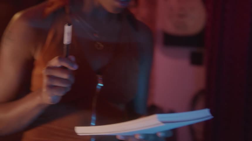 A woman is writing a song in a dimly lit music studio jotting down notes while another person plays a white electric guitar