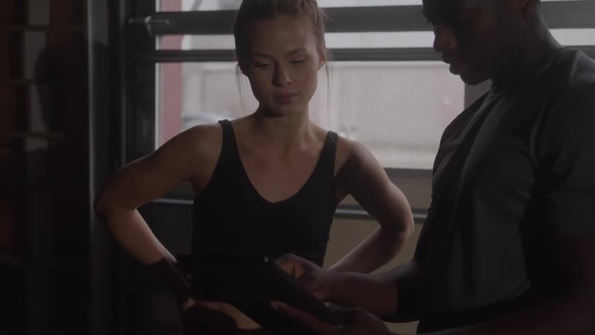 Young black man and young white woman standing indoors beside a window at midday the man is operating his mobile phone while the woman watches attentively