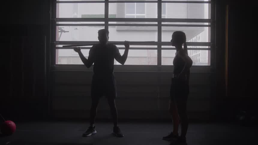 Man performs a squat with a barbell while a woman observes in the gym