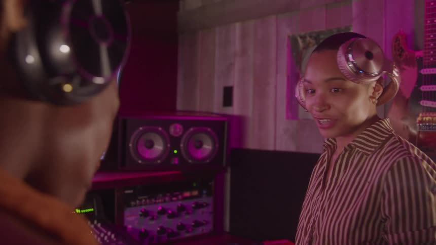 A young woman in a striped shirt and large headphones is talking and smiling at a man in a music studio with a guitar and audio equipment visible in the background