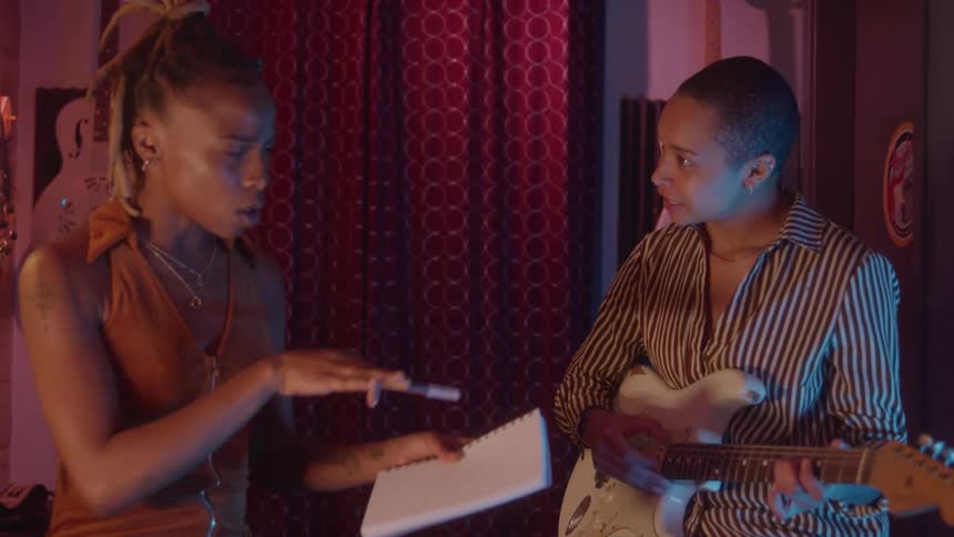 Two women rehearse in a studio one holding a notebook and pen while the other plays guitar