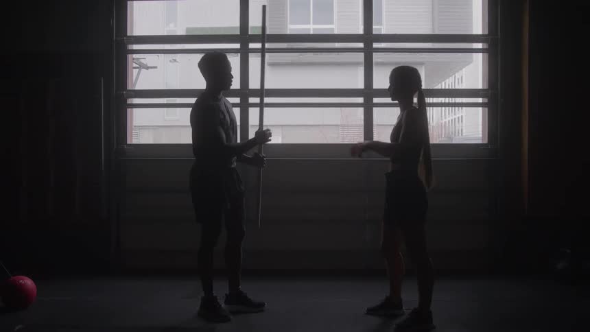 Silhouetted against a large window a man and woman train with a bar practicing squats and form