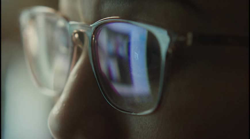 The video shows a closeup shot of a persons face focusing on their eyeglasses the glow of a screen likely a computer or tv is reflected in the lenses this suggests the person is looking at a digital device in a dimly lit room