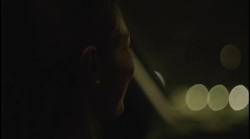 A mans profile is shown in a dark closeup shot with only the side of his face illuminated in the background there are blurry outoffocus lights possibly from a city street suggesting he is in a car at night