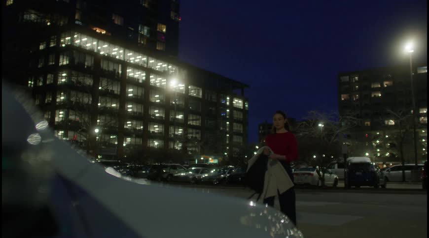 The video shows a woman in a red shirt walking through a parking lot at night she is holding a jacket and looking at a car with its hazard lights blinking in the background there are large buildings with many windows lit up