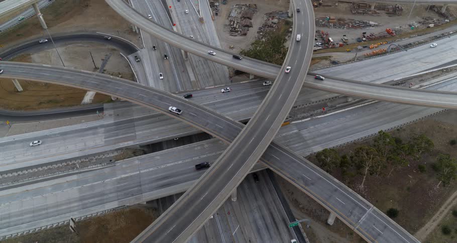The video shows a busy highway interchange from an aerial perspective cars and large trucks are seen traveling on the multilevel overpasses and the intersecting roads the light is flat and the sky is hazy suggesting an overcast day