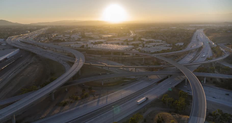 Aerial view of multiple interconnected overhead bridges leading in and out of an interstate highway bathed in the warm glow of an orange setting sun in the background