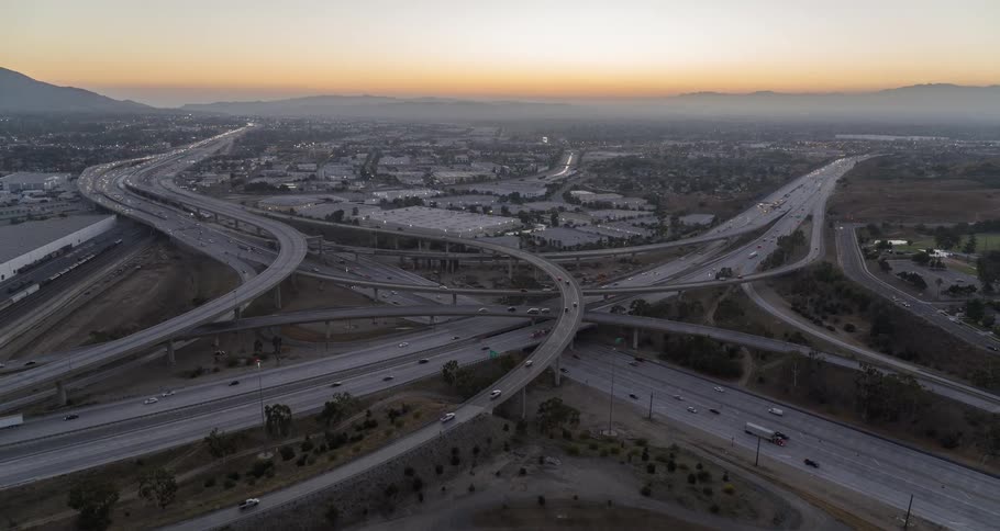 The video shows an aerial view of a massive highway interchange at dusk the complex network of overpasses and offramps is filled with cars and the surrounding city is visible in the background the sky has a faint orange glow from the setting sun