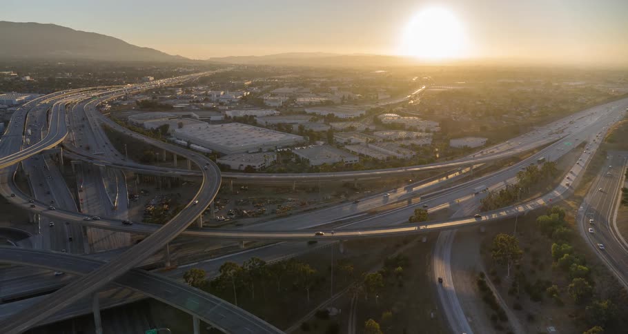 The video shows an aerial view of a complex highway interchange at sunset the sun is a bright light source on the horizon and cars with their headlights on can be seen moving along the roads