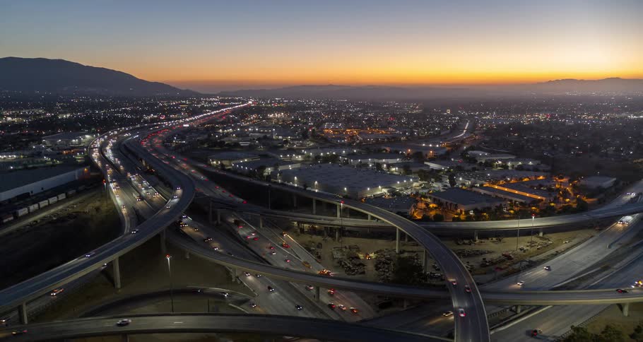 The video shows an aerial view of a city at dusk with the bright lights of buildings and a busy highway interchange illuminating the scene the sky is a gradient of orange and blue and mountains are visible in the background the traffic on the highway