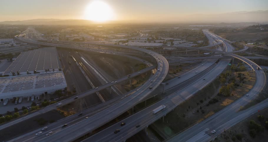 The video shows a wide aerial view of a multilevel highway interchange at sunset cars and large trucks are moving on the roads which are surrounded by commercial and industrial buildings the last light of the day illuminates the scene with a bright g