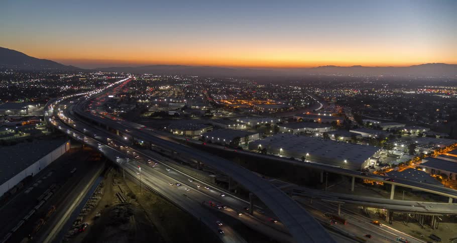 The video shows a busy highway interchange at sunset with a panoramic view of the surrounding city and mountains cars with their headlights on are moving along the multilevel overpasses as the sky changes from a deep orange to a darker twilight blue