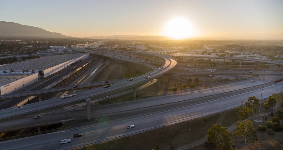 The video shows a sprawling highway interchange at sunset cars are seen traveling on multiple elevated overpasses and groundlevel lanes the sun is a bright orb on the horizon casting a golden light over the city and mountains in the distance