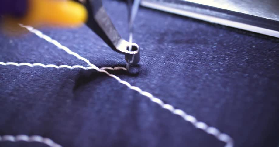 A sewing machine needle moves up and down while a thread creates a stitch on a piece of dark fabric