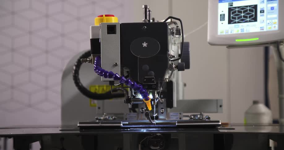 The video shows an automated industrial sewing machine a robotic arm guides a piece of fabric while the machines needle rapidly stitches a pattern a digital screen to the right displays a complex grid suggesting the machine is precisely following a pr