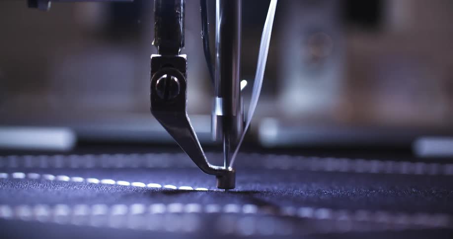 The video shows a closeup shot of a sewing machine needle the needle moves up and down rapidly creating a series of white stitches on a dark blue fabric the lighting highlights the needle and the stitches