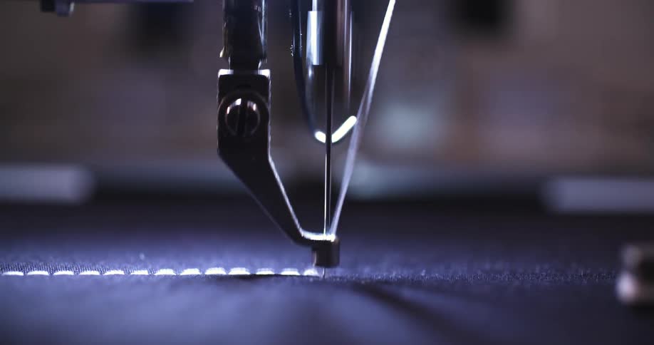 A sewing machine needle moves up and down while a thread creates a stitch on a piece of dark fabric