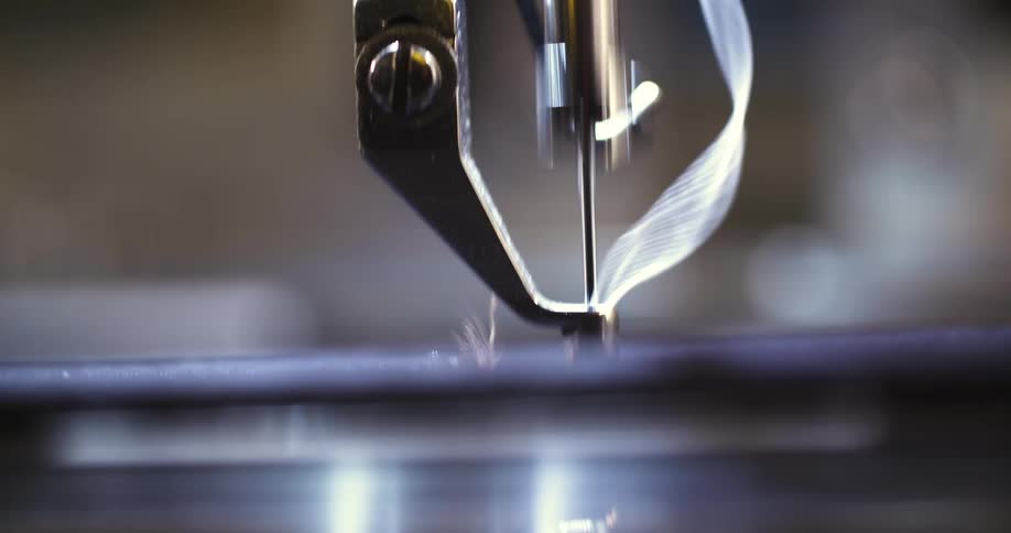 A sewing machine needle threaded with white thread moves precisely up and down beneath a metal presser foot stitching fabric securely in place