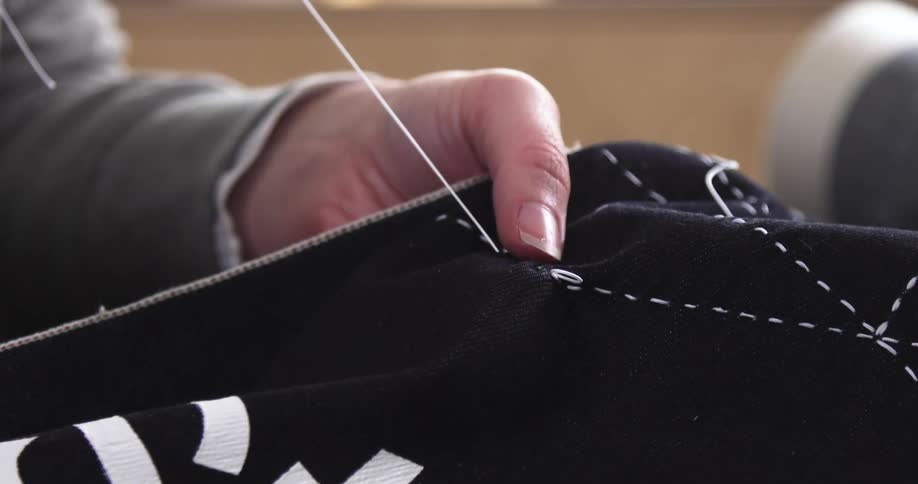 A person handstitches white thread through dark fabric carefully pulling it tight along the seam