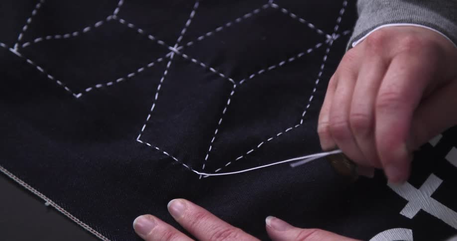 Hands carefully pull white thread through dark fabric patterned with stitched geometric lines tightening each handsewn stitch