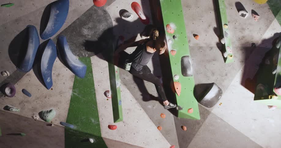A wide view from an angle shows a fit woman wearing a black tank top and leggings as she grips the gray green and orange handholds on a rock climbing wall