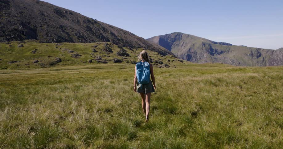 The video shows a wide shot of a woman with a blue backpack walking away from the camera through a grassy mountainous landscape the scene is bright and sunny highlighting the vast open field and the imposing rocky mountains in the background she is