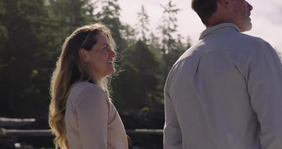 The video shows a man and a woman standing in a forested area with the woman on the left and her back to the camera the mans face is obscured but the woman turns to the side and smiles raising her hand to point at something out of frame the warm lig