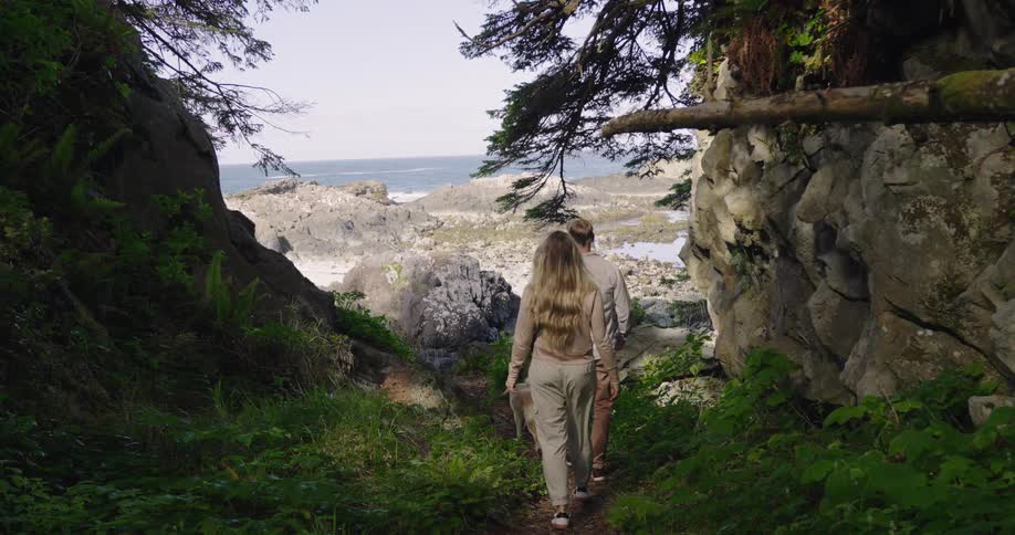 A couple with a dog walk down a lush rocky path framed by cliffs and trees with the ocean visible ahead