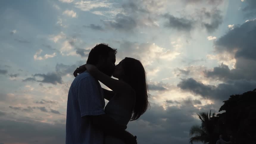 Silhouetted couple hug and kiss by palm trees as the sun sets