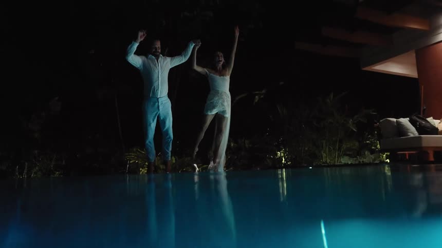 A young couple jump into a pool together at night while holding hands