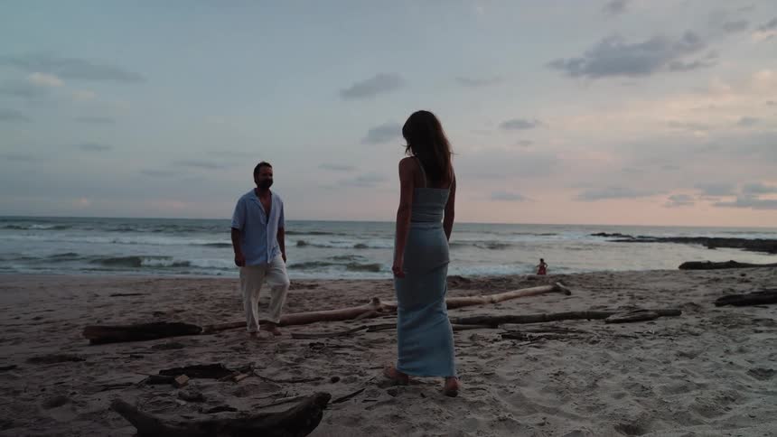 A woman in a long dress walks barefoot along the beach at sunset as a man approaches near the shoreline