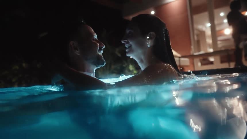 A couple partially submerged in a brightly lit swimming pool at night share an intimate moment and a kiss