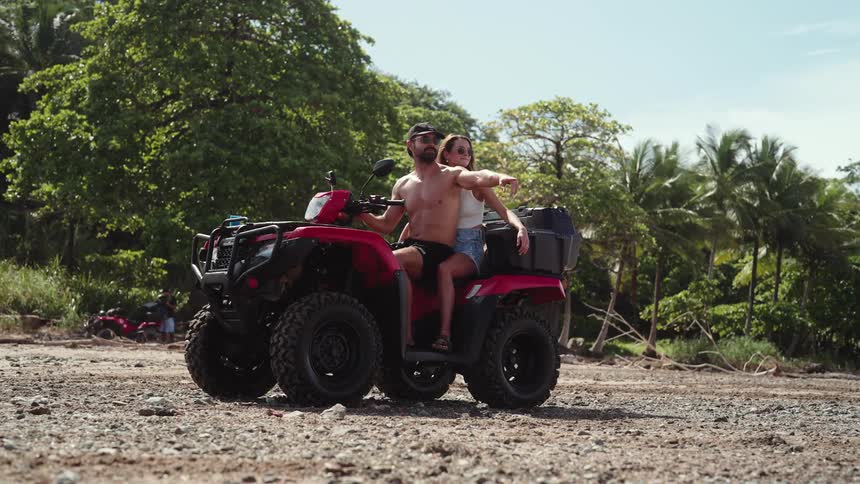 Couple riding a red atv through the woods under the midday sun with the shirtless young man wearing a hat and shorts pointing into the distance as the woman rides behind him
