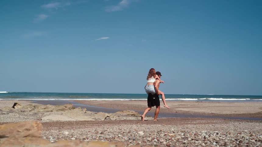 The video shows a wide shot of a man walking along a beach with a woman on his back in a piggyback ride they are moving from left to right across a wide expanse of sand with the bright blue ocean and sky behind them their playful interaction suggests a