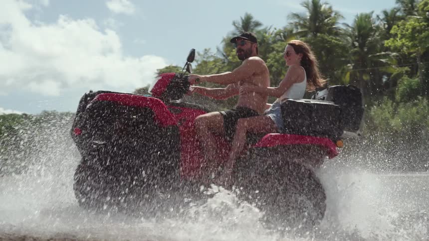 The video shows a man and a woman on a red quad bike the vehicle emerges from a large splash of water creating a dramatic wake the couple rides away from the splash looking happy and carefree as they enjoy their adventurous ride on a sunny day