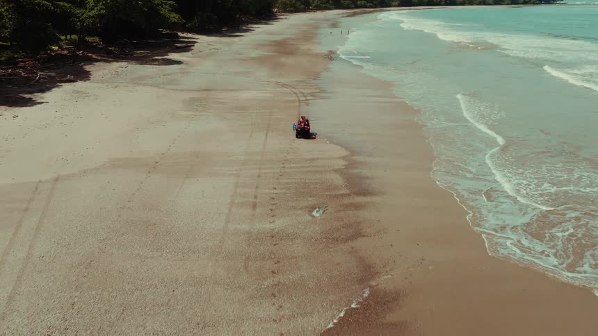 An atv carrying three people drives along a wide sandy beach with gentle waves lapping the shore and lush green trees bordering the far side