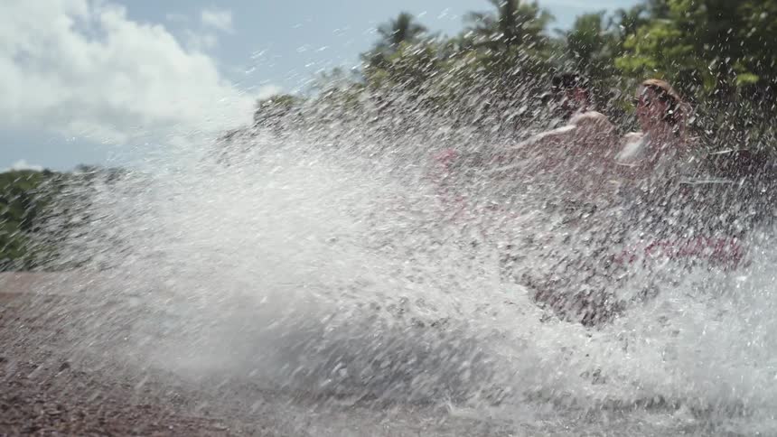 A man and woman are riding a red quad bike on a beach as they drive through the shallow water they create a large splash