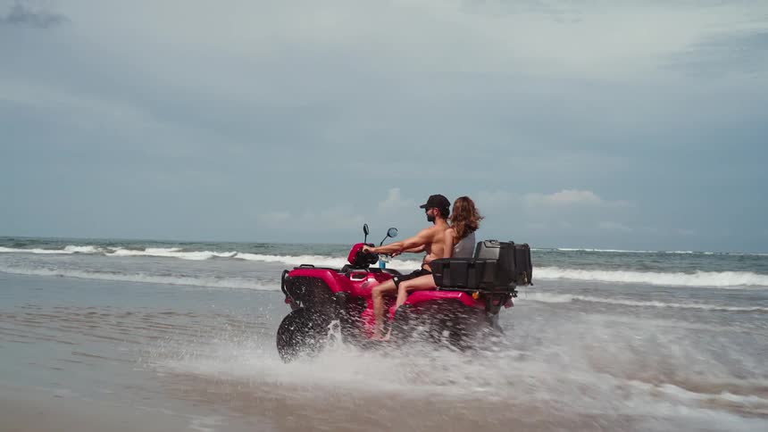 Couple riding a red atv through shallow beach water at midday with the shirtless young man wearing a hat and shorts as the woman rides behind him and the wind blows her long brown hair behind her