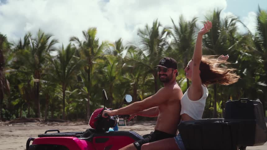 Couple rides a red atv along the beach with palm trees in the background