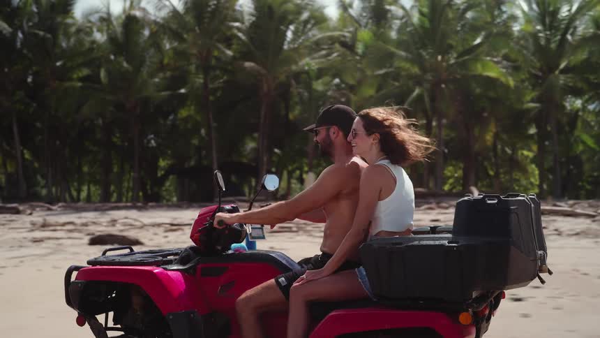 A man and woman ride together on a red atv along the beach wind in their hair with palm trees behind them