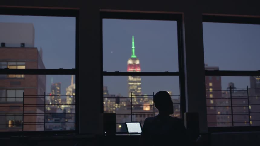 A man swivels in his desk chair in front of large windows
