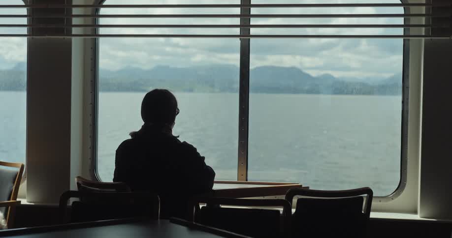 The silhouette of a person sits at a table and looks out a window at a body of water and mountains